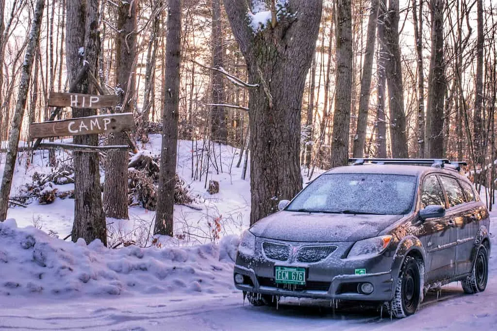 a grey Pontiac Vibe parked in the snow next to a Hipcamp sign while on a Vermont glamping trip.