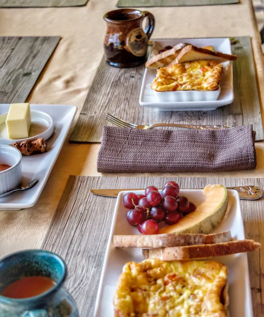 An outdoor breakfast table setting at Moose Meadow Lodge and Treehouse, featuring omelettes, toast, fresh fruit, and coffee.
