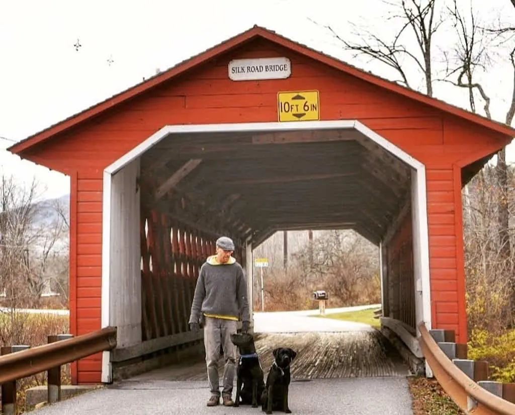 A man stands with two black labs in front of the Silk Road Covered Bridge in Bennington, Vermont.