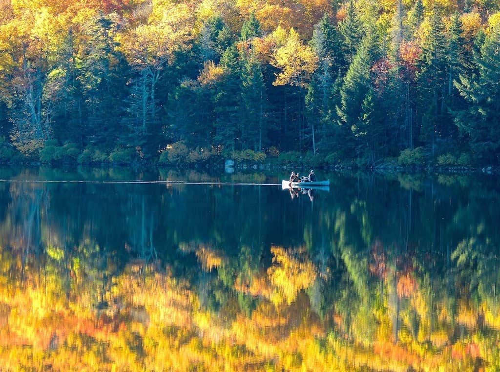 A canoe slices through calm water during fall foliage season on Grout Pond in Vermont.