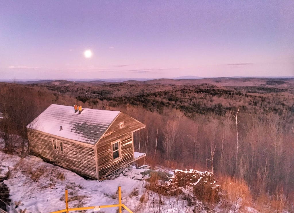 The winter view from Hogback Mountain in Wilmington, Vermont featuring an abandoned cabin, snow, and the full moon.