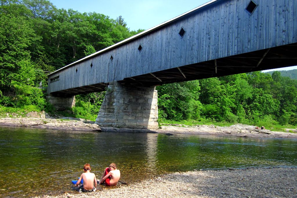 Two kids sit on the edge of the river under the West Dummerston Covered Bridge in Vermont.
