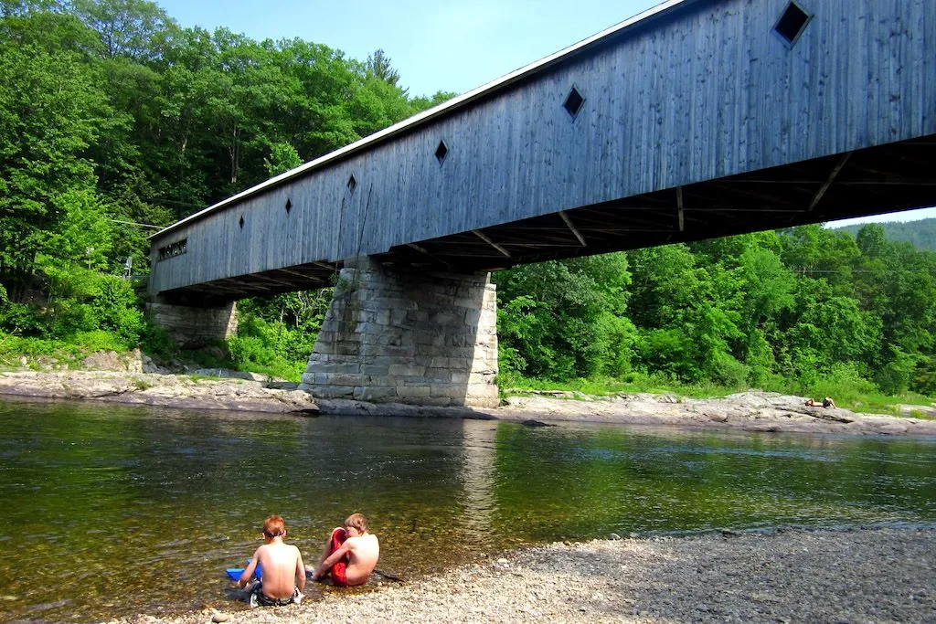 Two kids sit on the edge of the river under the West Dummerston Covered Bridge in Vermont.