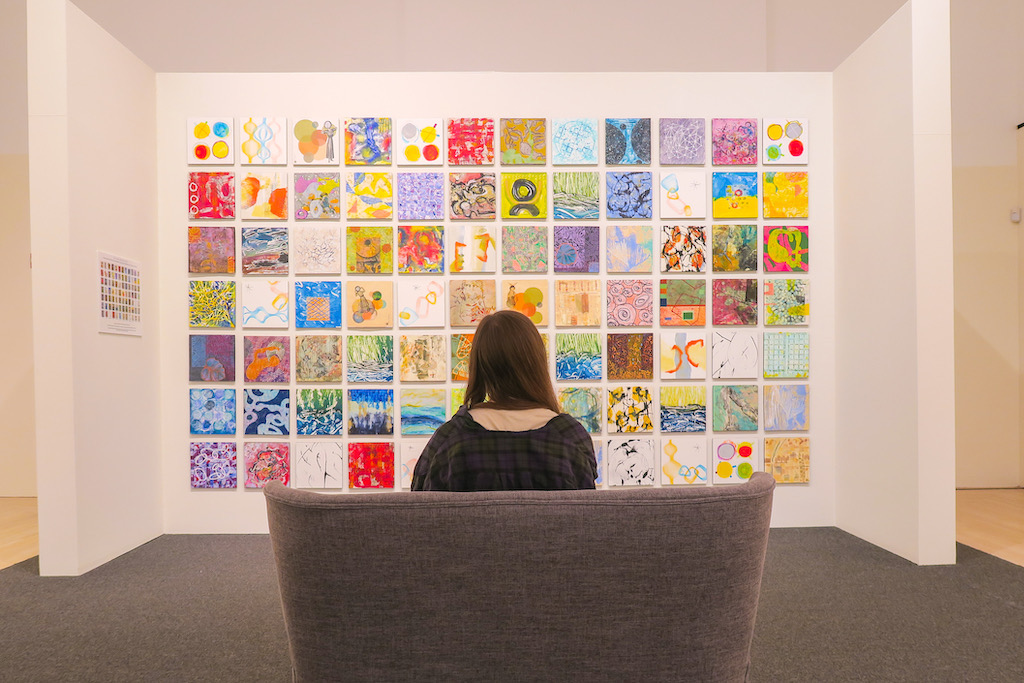 A woman sits with her back to the camera looking at a wall of colorful paintings.