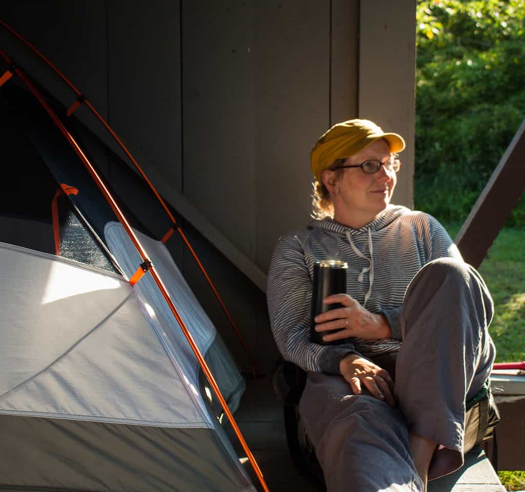 Tara sitting next to her tent holding a cup of coffee in a travel mug.
