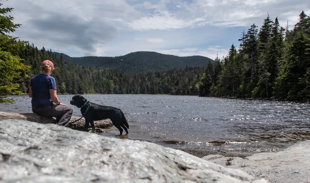 Tara sits on a rock next to Sterling Pond while black lab, Malinda stands nearby. 